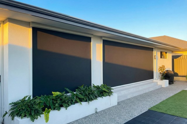 Two dark outdoor blinds lowered across the wall of an alfresco on a white rendered house, with ferns in a planter below